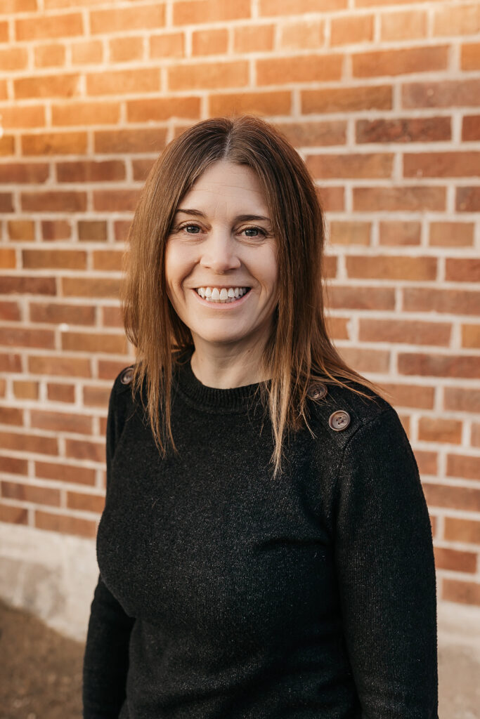 Maria Belmont smiles in front of a brick wall, wearing a black sweater.