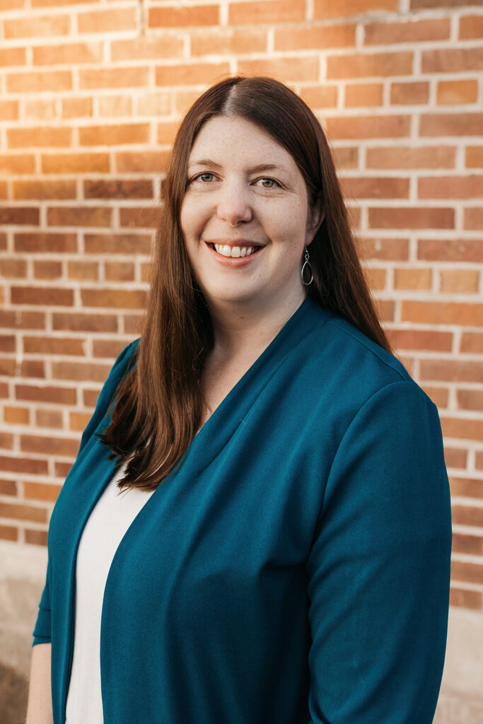 Maria Belmont smiling in front of a brick wall, wearing a teal blazer.
