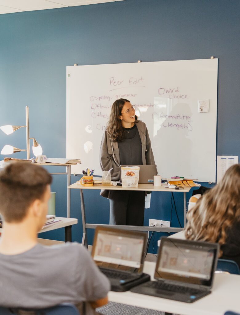 Teacher leading a peer edit session in a classroom with students and laptops. "Peer Edit" is written on the whiteboard.
