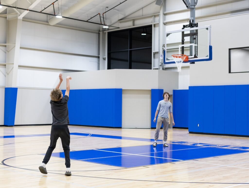 Two people playing basketball in a blue-walled indoor gym with wooden floors