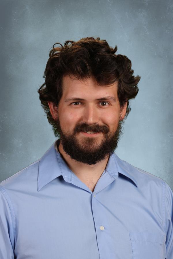Portrait of a man with dark curly hair and a beard, wearing a light blue collared shirt.