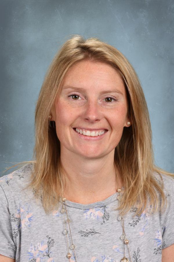 Portrait of a smiling blonde woman with fair skin and freckles, wearing a grey floral shirt and beaded necklace.