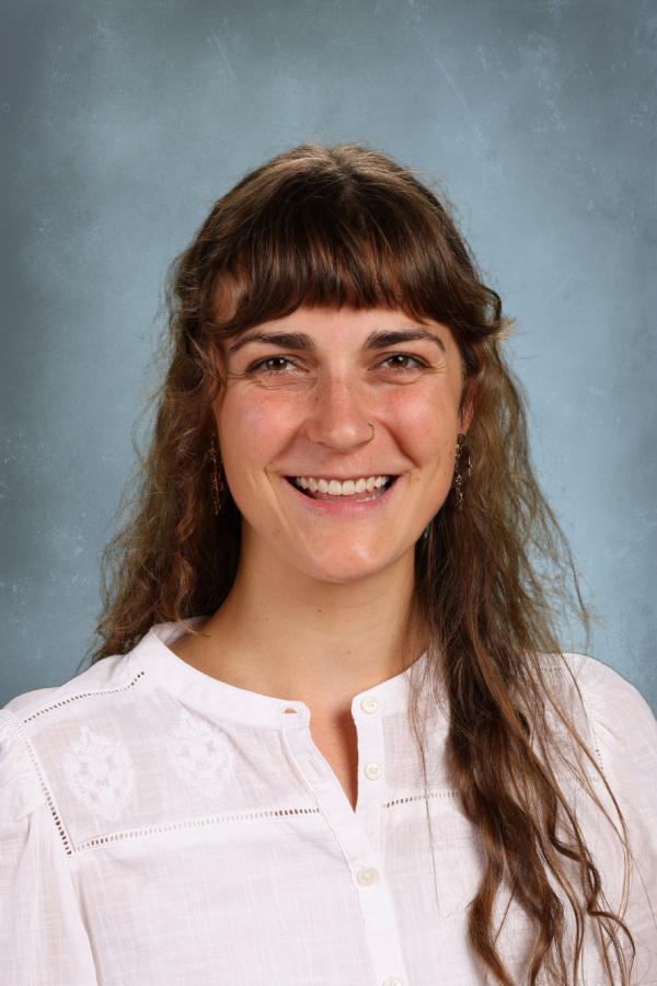Portrait of a smiling woman with long, wavy brown hair and bangs, wearing a white embroidered shirt.