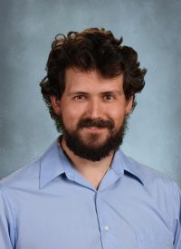 Portrait of a man with dark curly hair and a beard, wearing a light blue collared shirt.