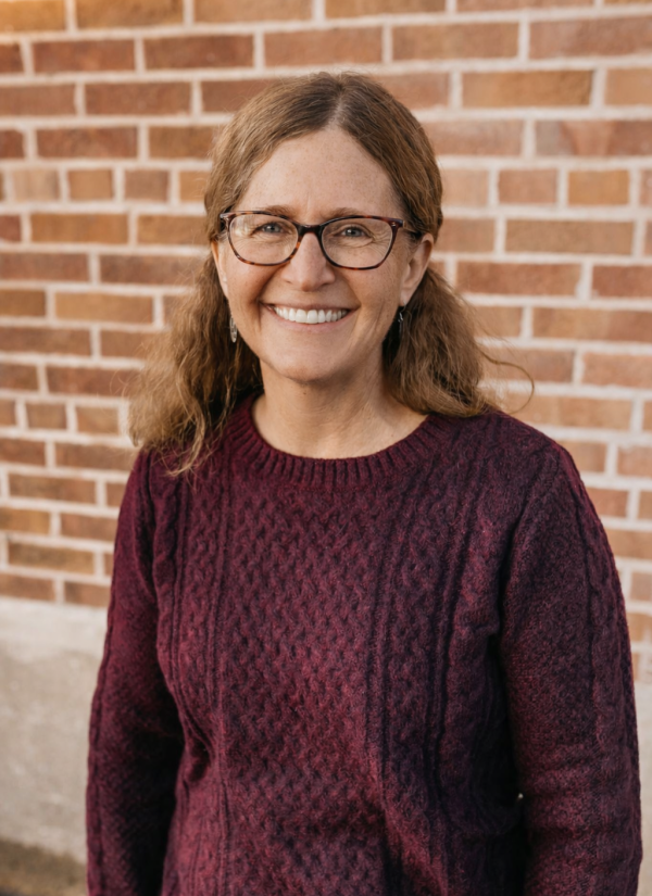 Faith Nadelhoffer smiling in front of a brick wall, wearing glasses and a maroon sweater.