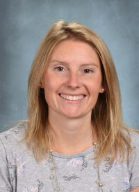 Portrait of a smiling blonde woman with fair skin and freckles, wearing a grey floral shirt and beaded necklace.