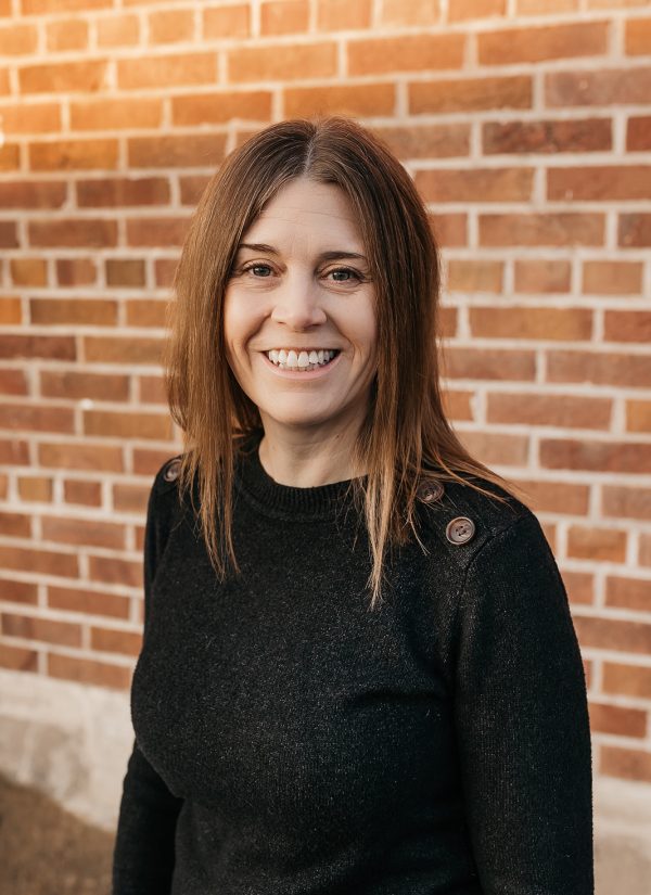Maria Belmont smiles in front of a brick wall, wearing a black sweater.