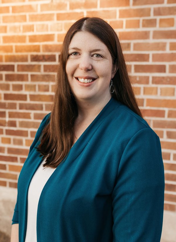 Maria Belmont smiling in front of a brick wall, wearing a teal blazer.