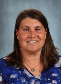 Portrait of a smiling woman with brown hair wearing a blue floral top and a heart necklace.