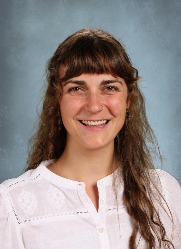 Portrait of a smiling woman with long, wavy brown hair and bangs, wearing a white embroidered shirt.
