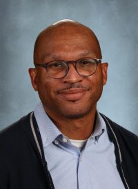 Portrait of a Black man with glasses and a light blue collared shirt.